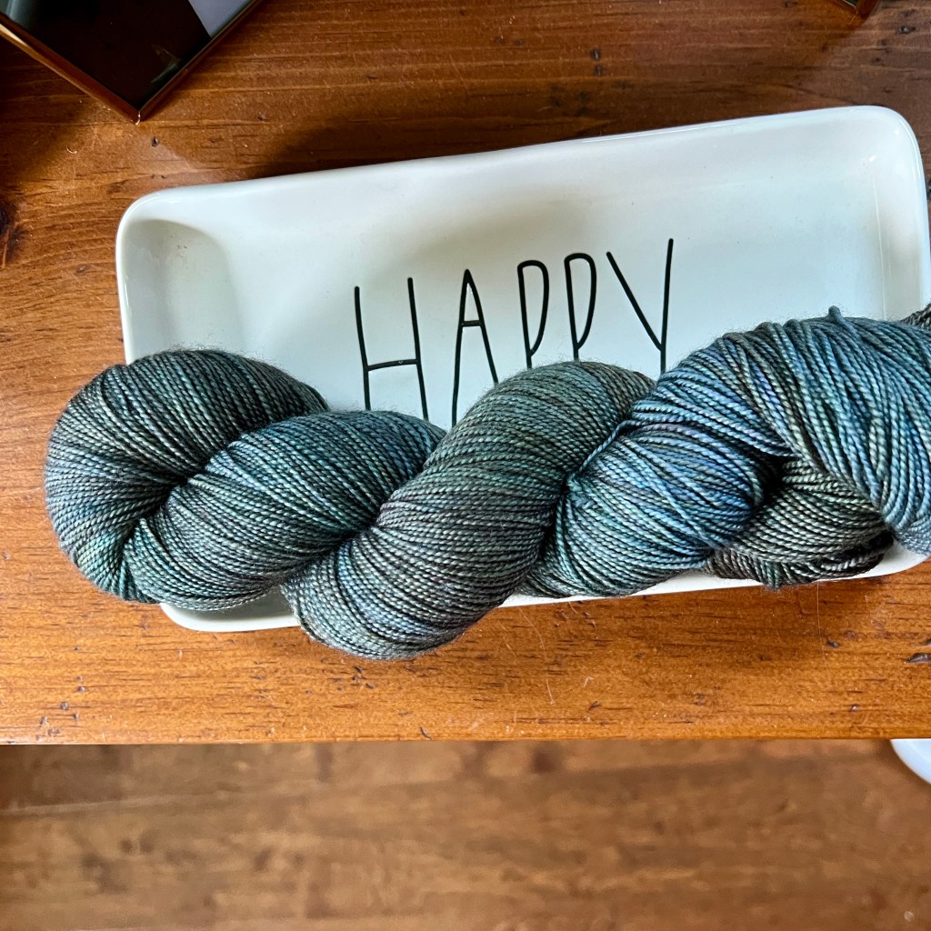 A skein of yarn in shades of blue and green placed on a white dish that has the word 'HAPPY' written on it.
