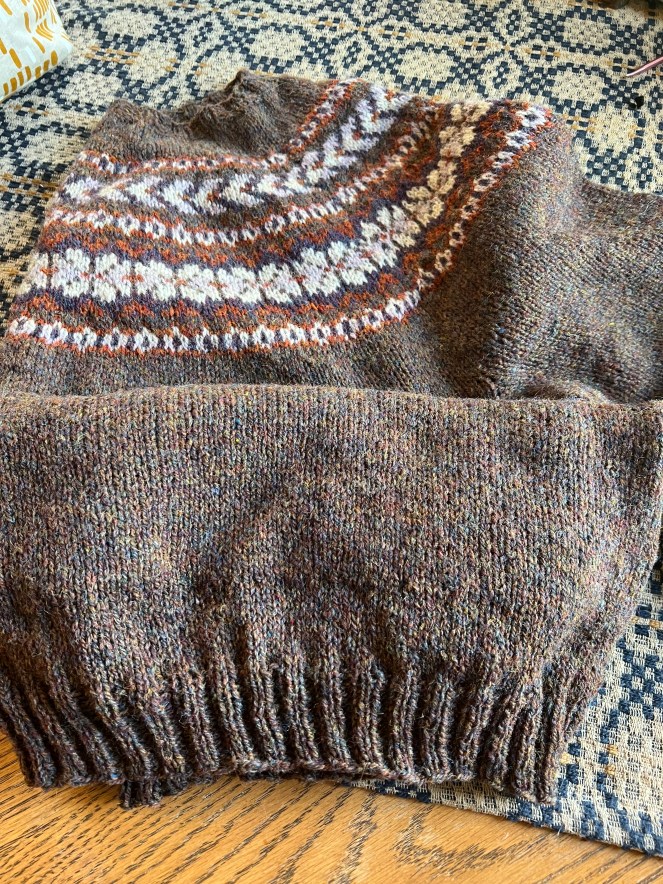 Close-up view of a hand-knit sweater showing a patterned yoke in browns and cream with a brown ribbed cuff, placed on a wooden surface.