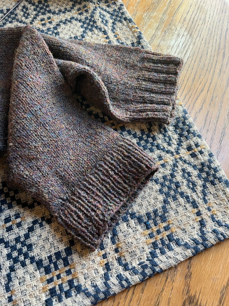 Close-up of a knitted sweater sleeve cuff in textured brown wool resting on a patterned fabric surface.
