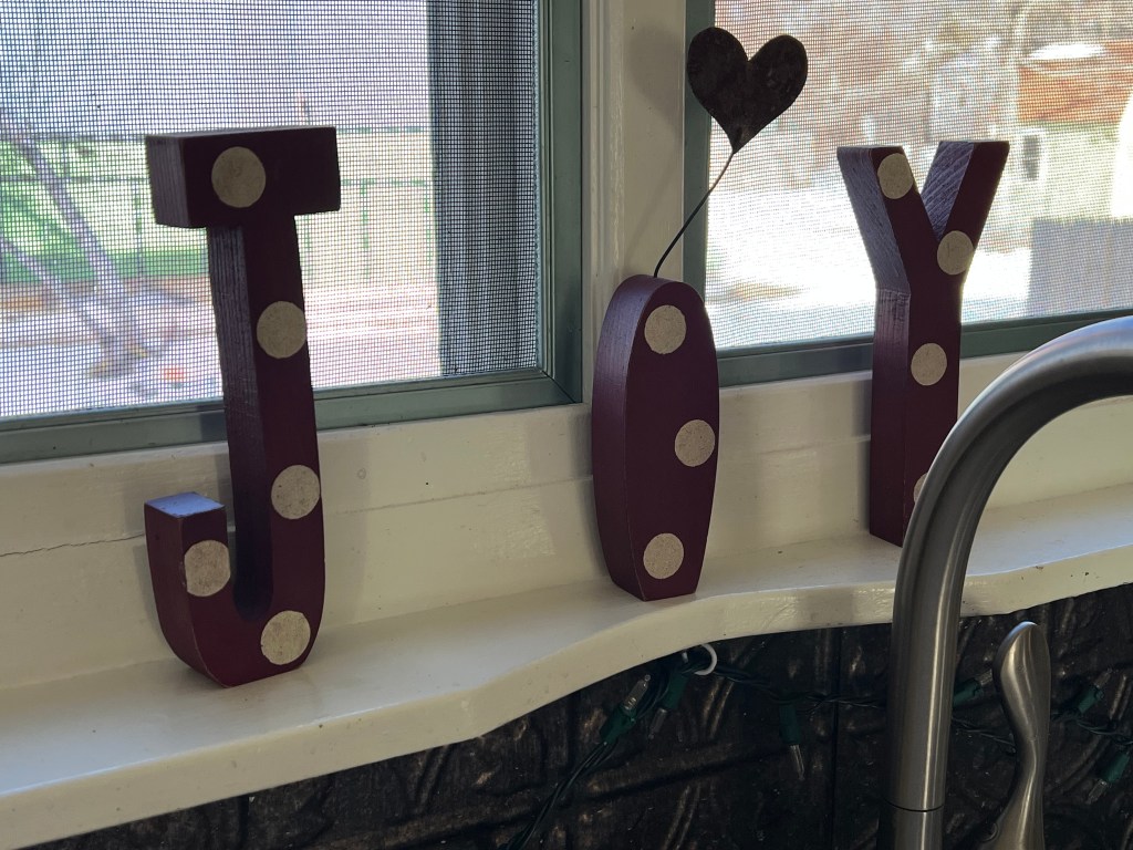 Decorative letters spelling 'JOY' in maroon color with white dots, placed on a windowsill with a small heart decoration.