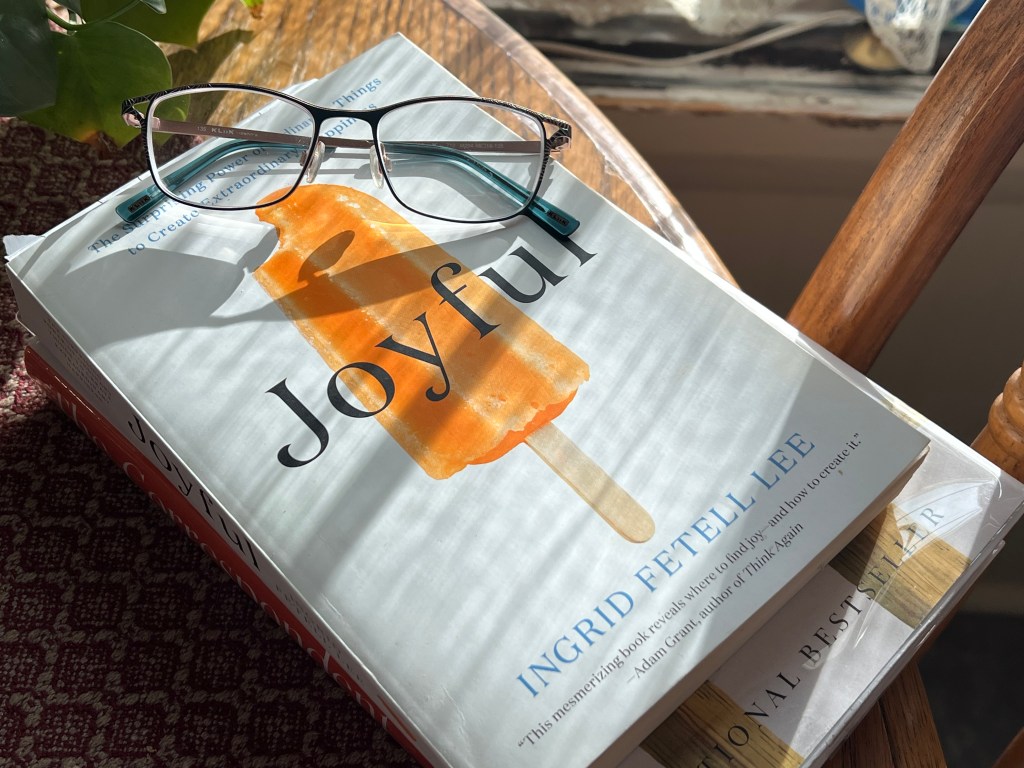 A book titled 'Joyful' by Ingrid Fetell Lee resting on a table, with a pair of glasses placed on top. The book features a bright cover with an image of a popsicle, surrounded by warm sunlight and a hint of greenery in the background.