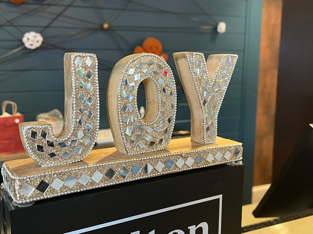 Decorative wooden letters spelling 'JOY' adorned with mosaic patterns, set against a dark background with a hotel reception area visible.